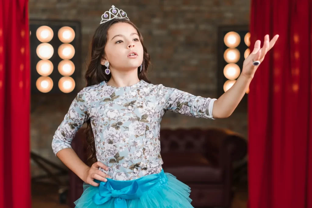 Child wearing a tiara performing between red stage curtains